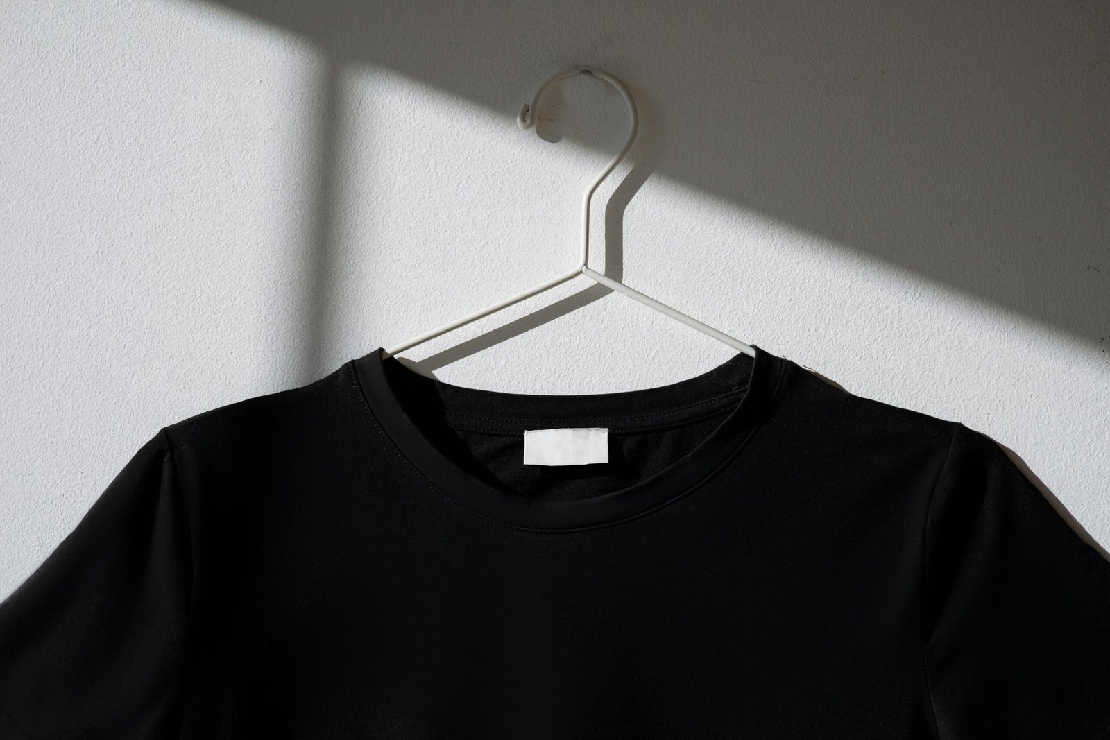 Black t-shirt hanging on a white hanger against a plain wall with a dramatic shadow effect.