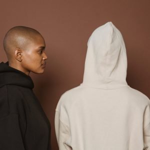 Unemotional bald African American female in oversize hoodie standing near person in similar outfit against brown background
